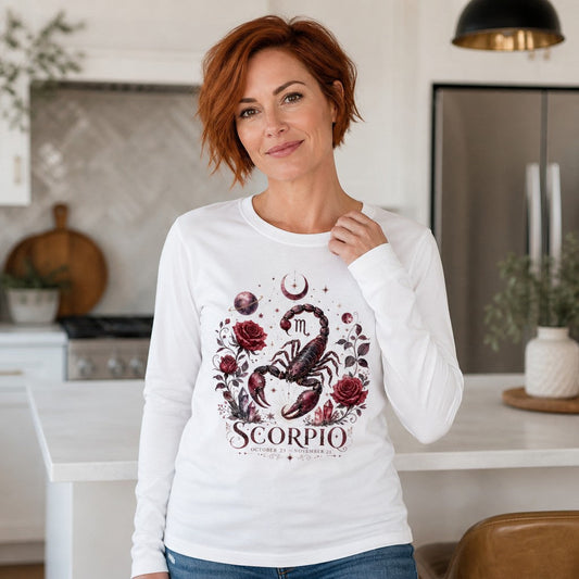 Woman wearing white Bella Canvas 3501 long sleeve shirt featuring a hand-painted Scorpio scorpion with deep red roses, amethyst crystals, crescent moons, and celestial details in vintage botanical cottagecore style, standing in a cozy kitchen, ideal for astrology and cottagecore lovers, by Crossroads Dandelion