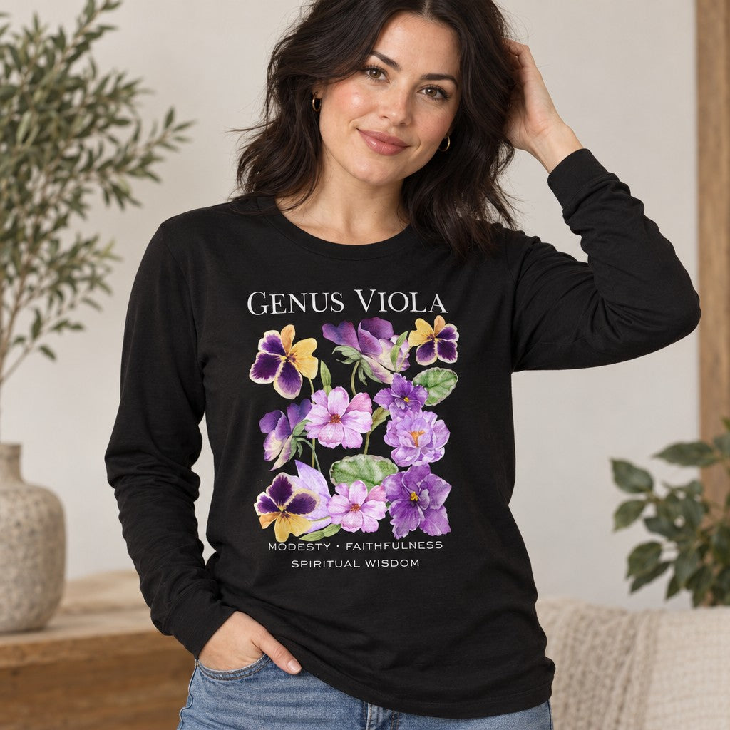 Woman wearing a black Bella Canvas 3501 long sleeve shirt featuring a watercolor bouquet of violets, pansies, and heart-shaped leaves with hand-lettered botanical symbolism for modesty, faithfulness, and spiritual wisdom in a vintage cottagecore style, perfect for botanical art lovers, indoor setting, by Crossroads Dandelion