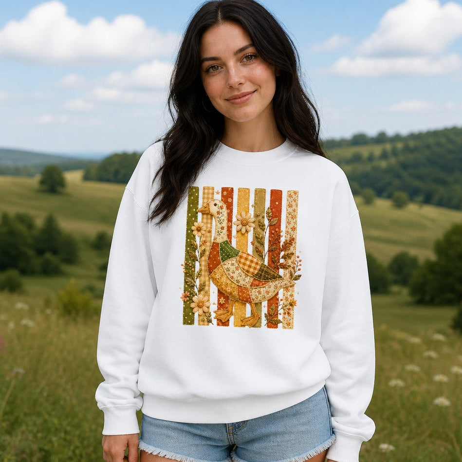 Woman wearing a white Comfort Colors 1566 sweatshirt featuring a whimsical patchwork goose with floral, gingham, and polka-dot patterns, vintage folk art style with embroidered wildflowers on stitched slats, set in a rural outdoor scene, cozy cottagecore autumn garden lovers, by Crossroads Dandelion