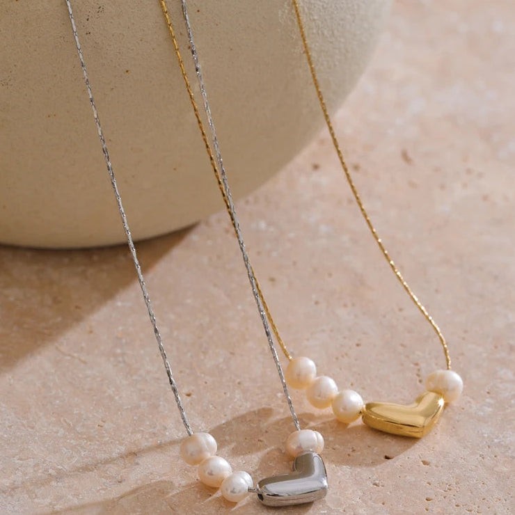 Gold and silver pearl heart necklaces draped over a cream vase onto warm stone