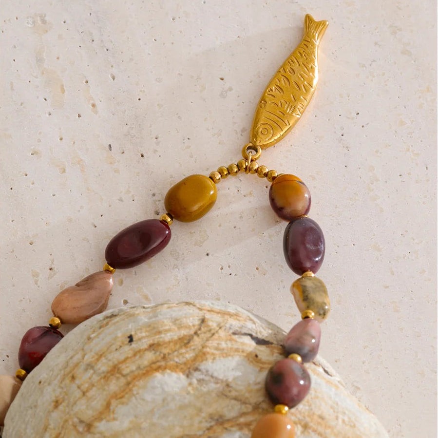Close-up of a gold fish pendant and natural stone beads resting on a seashell