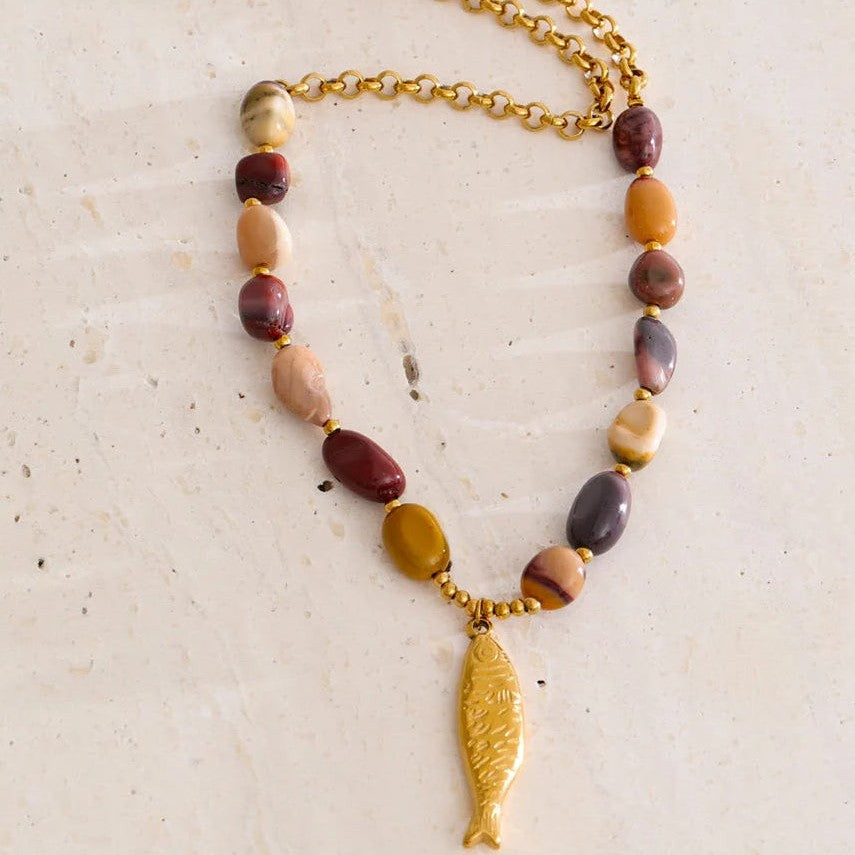 Gold fish pendant necklace with stone beads and rolo chain laid on travertine