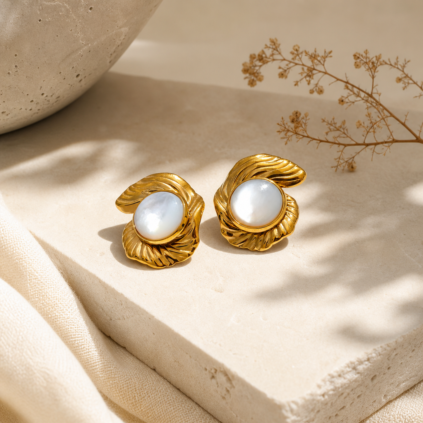 Golden leaf-shaped stud earrings with iridescent pearl centers displayed on textured beige stone, elegant jewelry product photography, by Crossroads Dandelion