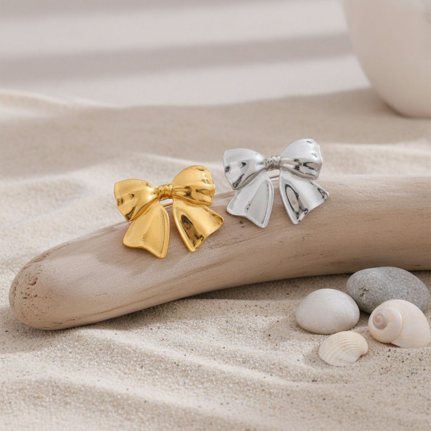 Gold and silver bow brooches displayed on sand with shells, by Crossroads Dandelion