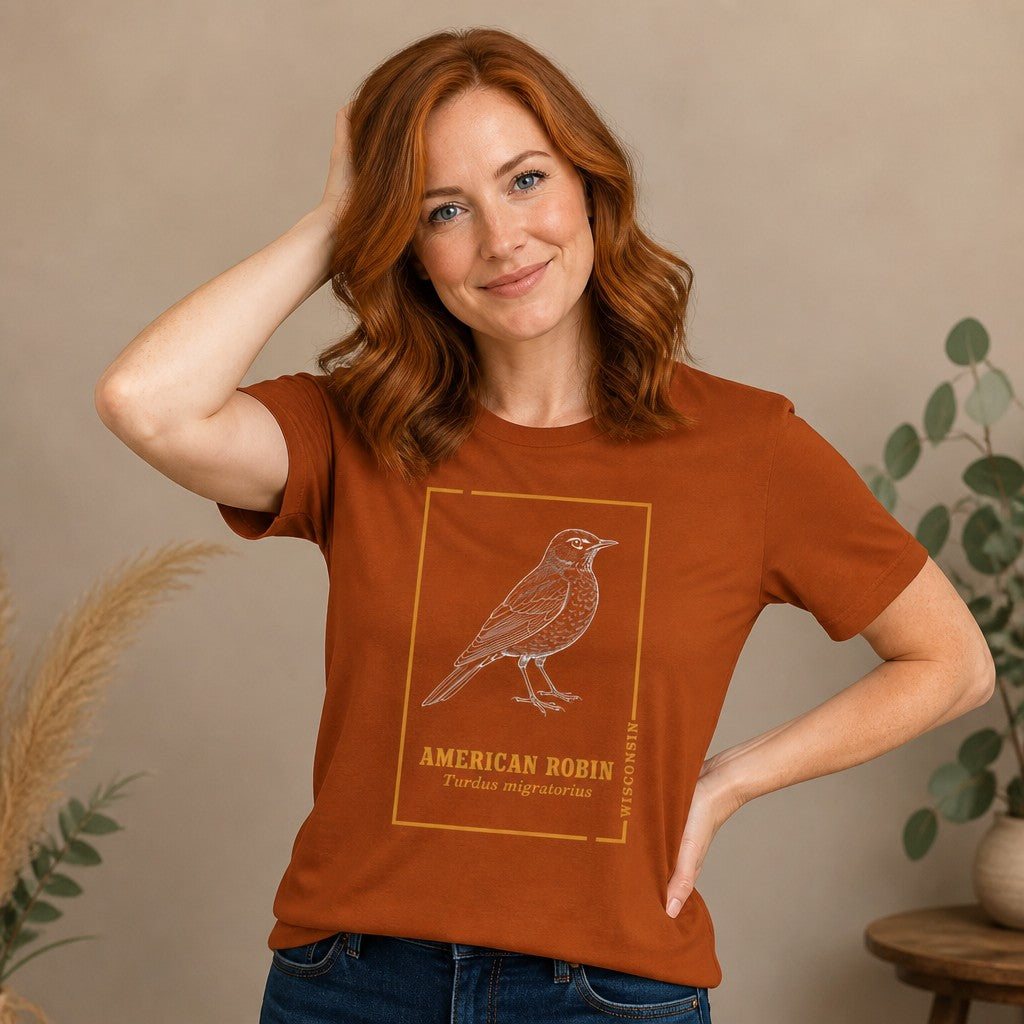 Woman wearing an autumn-colored Bella Canvas 3001 t-shirt featuring a detailed line art illustration of an American Robin with vintage botanical accents, styled for cottagecore and birdwatching enthusiasts, indoor scene, by Crossroads Dandelion