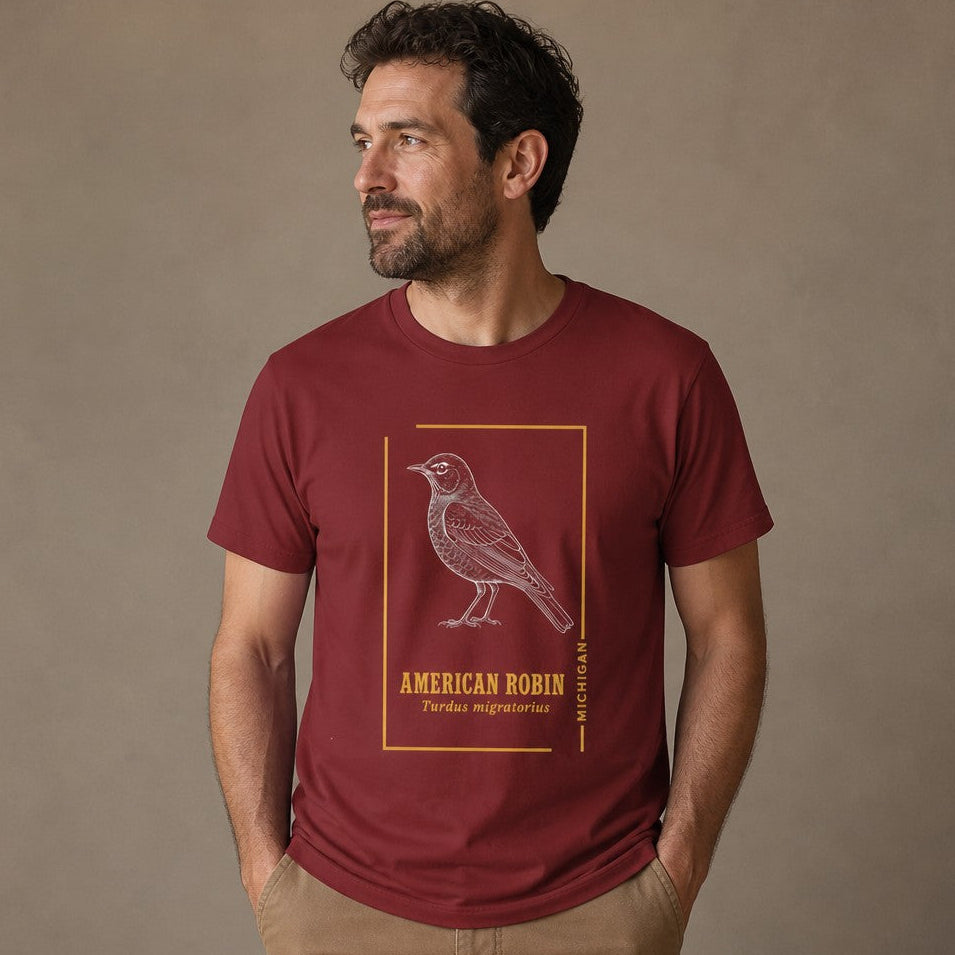 Man wearing a cardinal red Bella Canvas 3001 t-shirt featuring an intricate white line art illustration of an American robin in a vintage nature field-guide style, minimalist gold border, perfect for birdwatchers, naturalists, and cottagecore enthusiasts, by Crossroads Dandelion