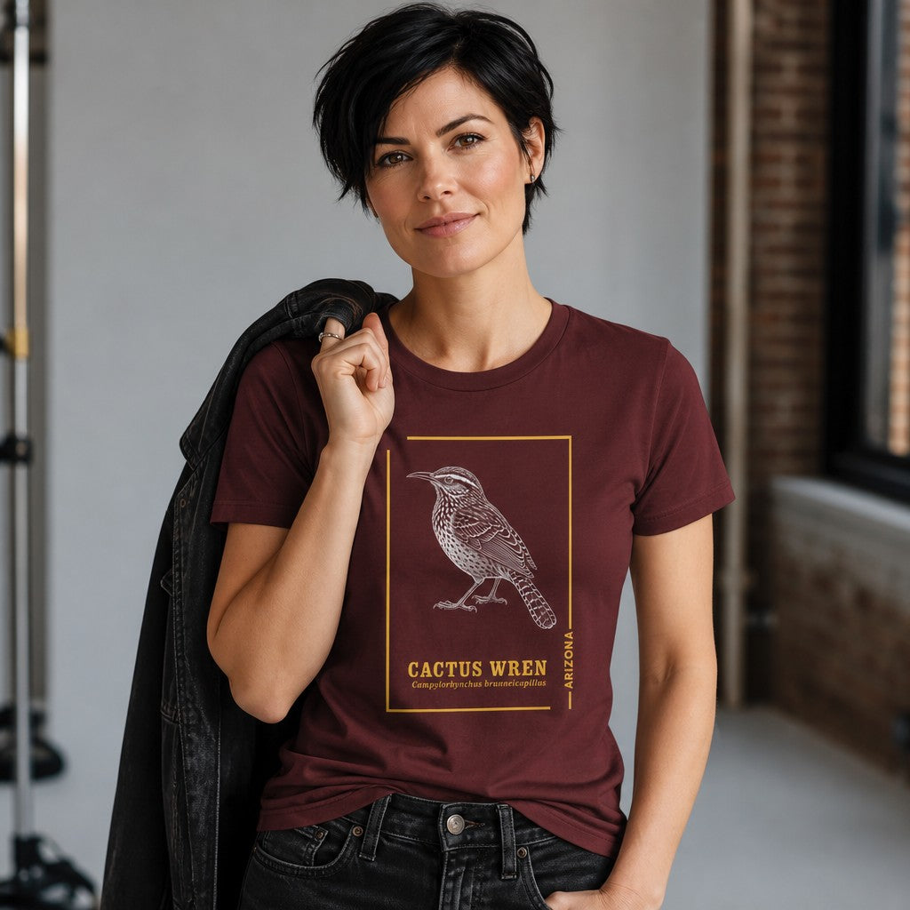 Woman wearing a maroon Bella Canvas 3001 t-shirt featuring a vintage line art illustration of the Cactus Wren, Arizona state bird, with golden frame and botanical accents, loft setting, perfect for lovers of ornithology, desert flora, and Southwest style, by Crossroads Dandelion