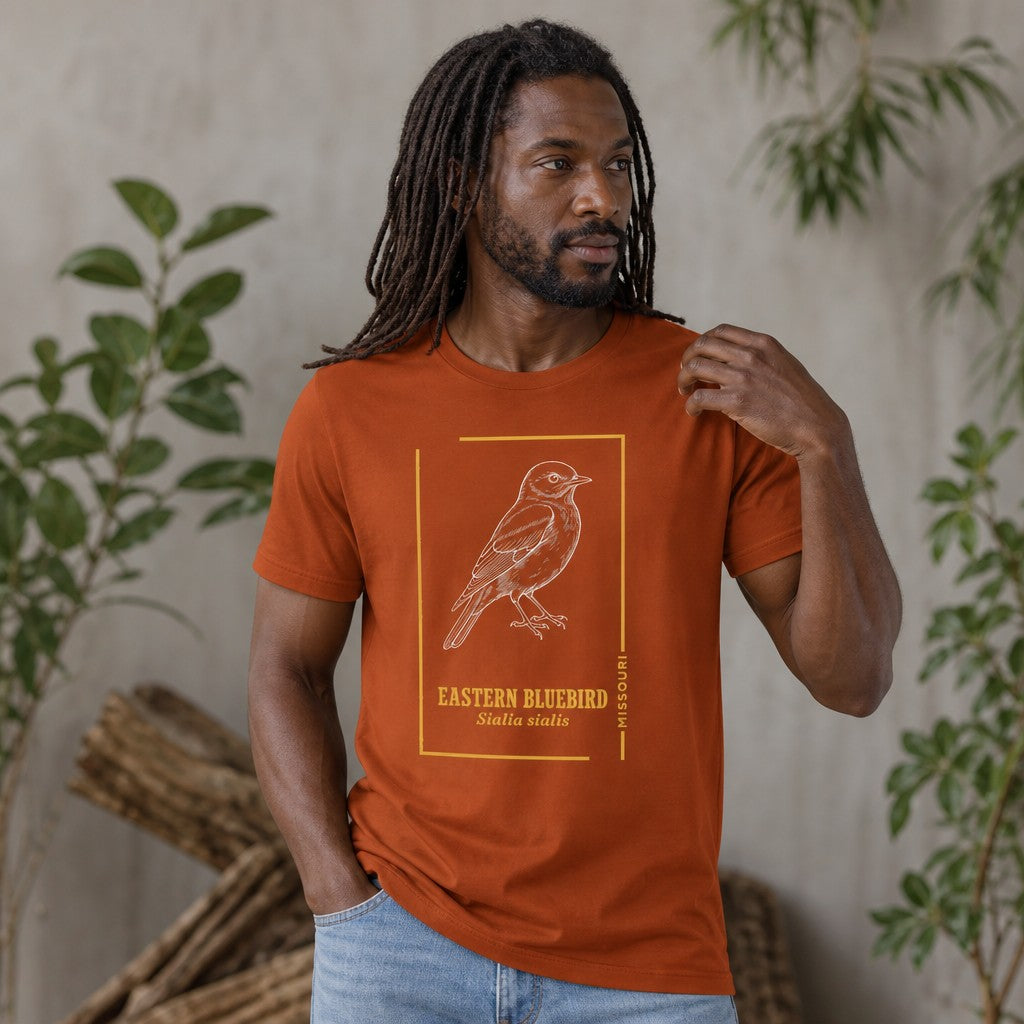 Man wearing autumn orange Bella Canvas 3001 t-shirt featuring a vintage botanical line art illustration of an Eastern Bluebird with golden accents and scientific name, styled for birdwatchers and nature lovers, indoor botanical setting, by Crossroads Dandelion