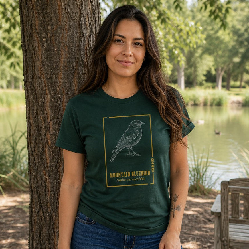 Woman wearing a forest green Bella Canvas 3001 t-shirt with a vintage-inspired line art illustration of a Mountain Bluebird framed by golden accents and scientific name, nature park scene, ideal for birdwatchers and nature lovers, by Crossroads Dandelion