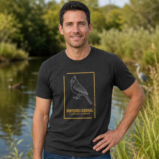 Man in dark grey heather Bella Canvas 3001 t-shirt featuring a hand-drawn vintage-style line art illustration of a Northern Cardinal framed in gold, inspired by botanical field journals, perfect for birdwatchers and cottagecore naturalists, standing by a pond in nature, by Crossroads Dandelion