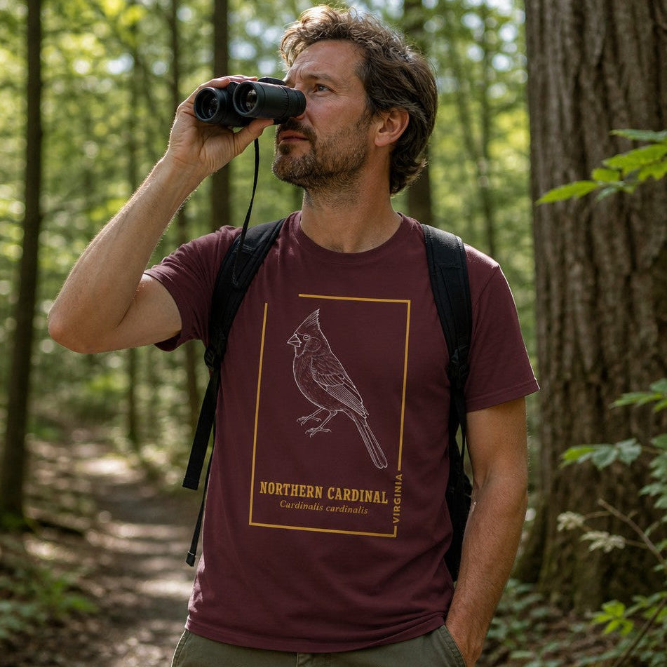 Man wearing a maroon Bella Canvas 3001 t-shirt with a vintage white line art illustration of a northern cardinal, accented by gold framing and serif text, cottagecore style, birdwatcher hiking in a lush forest, perfect for nature lovers and birding enthusiasts, by Crossroads Dandelion
