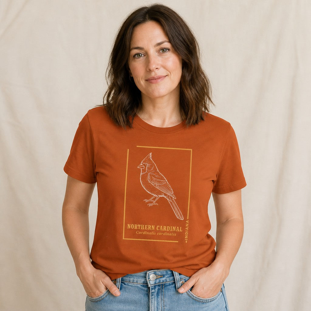 Woman wearing an autumn orange Bella Canvas 3001 t-shirt featuring a vintage-inspired line art illustration of a Northern Cardinal perched, accented with field guide typography and state callout, ideal for wildlife art and birdwatching enthusiasts, relaxed indoor setting, by Crossroads Dandelion