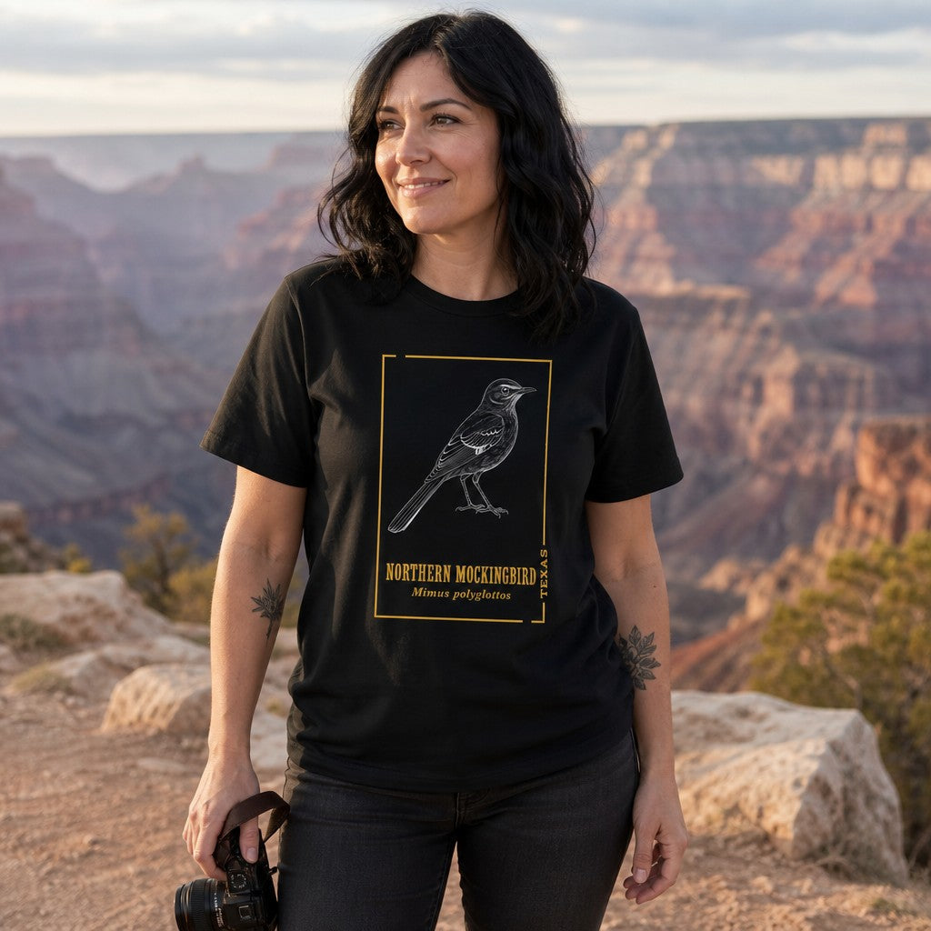 Woman wearing black t-shirt with minimalist line art northern mockingbird design and yellow text, standing at the Grand Canyon, birdwatcher apparel for women, by Crossroads Dandelion