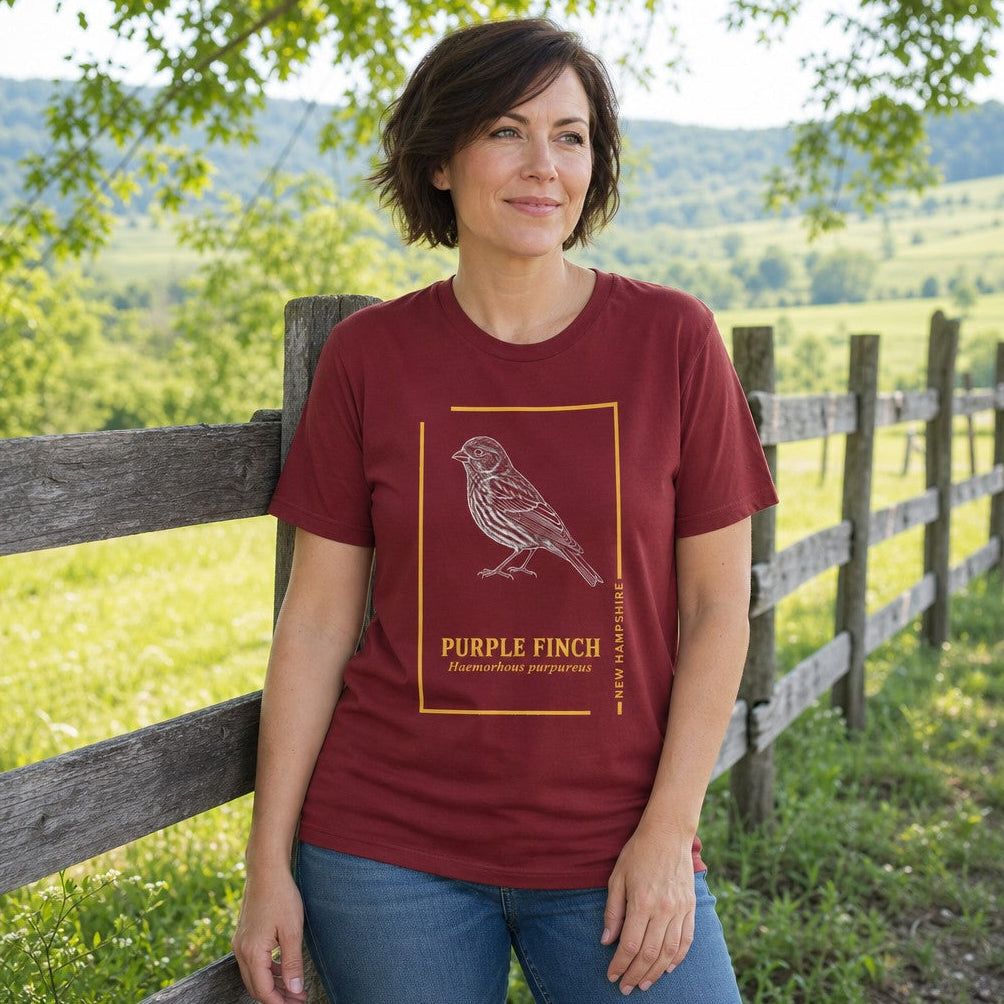 Woman in cardinal red t-shirt featuring a line art purple finch bird illustration and botanical text, standing by a wooden fence in a rural countryside setting, perfect for nature and bird lovers, by Crossroads Dandelion
