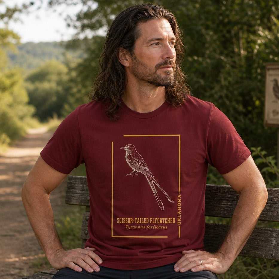 Man wearing cardinal Bella Canvas 3001 t-shirt featuring a vintage-inspired line drawing of a scissor-tailed flycatcher within a golden frame, naturalist botanical bird illustration, seated on an outdoor bench along a wooded trail, perfect for bird lovers and nature enthusiasts, by Crossroads Dandelion