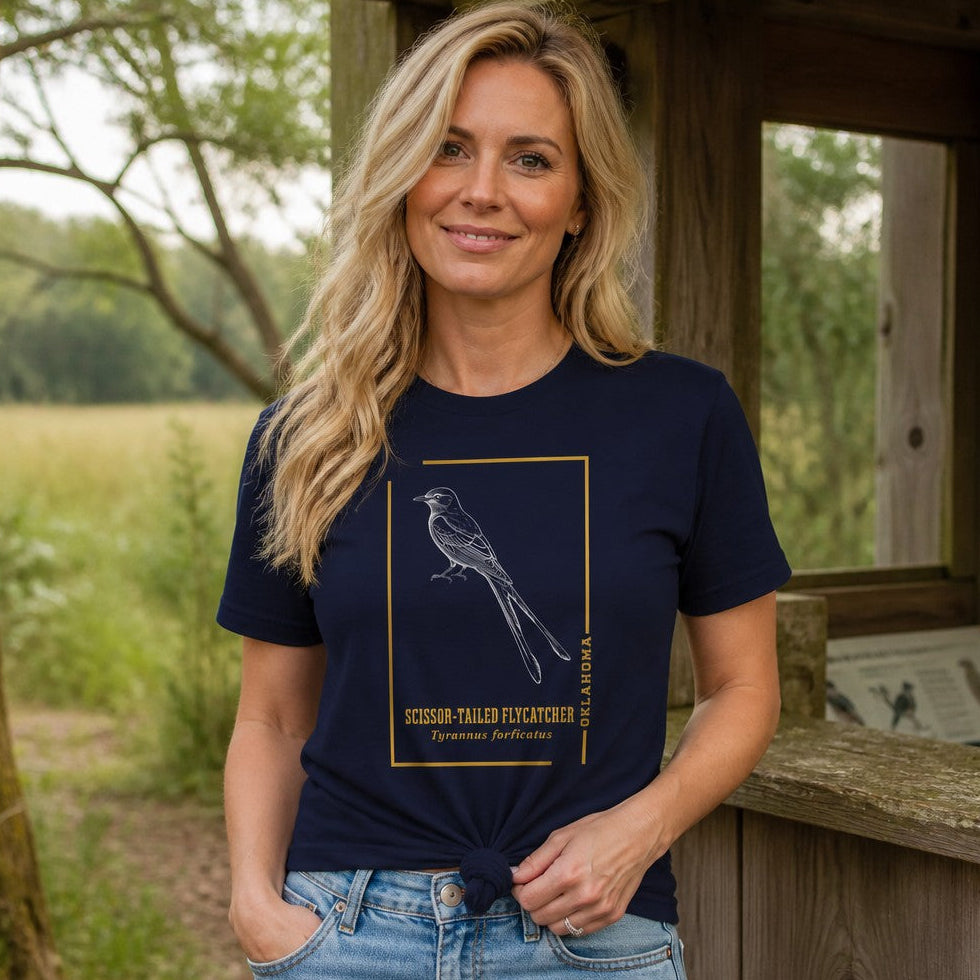 Woman wearing a navy Bella Canvas 3001 t-shirt featuring a vintage-inspired line art drawing of a scissor-tailed flycatcher inside a golden frame, set outdoors on a rustic porch with prairie grass in the background, perfect for bird lovers, gardeners, and nature enthusiasts, by Crossroads Dandelion