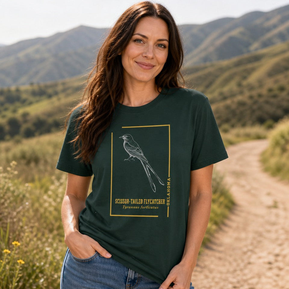 Woman wearing a forest green t-shirt with a line art illustration of a scissor-tailed flycatcher and scientific name, standing on a nature trail with mountains in the background, perfect for birdwatchers and nature lovers, by Crossroads Dandelion