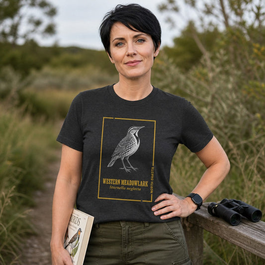 Woman wearing dark grey heather Bella Canvas 3001 t-shirt with vintage-inspired line art illustration of a Western Meadowlark framed in gold, standing on a nature trail while birdwatching, ideal for classic field guide art lovers, by Crossroads Dandelion