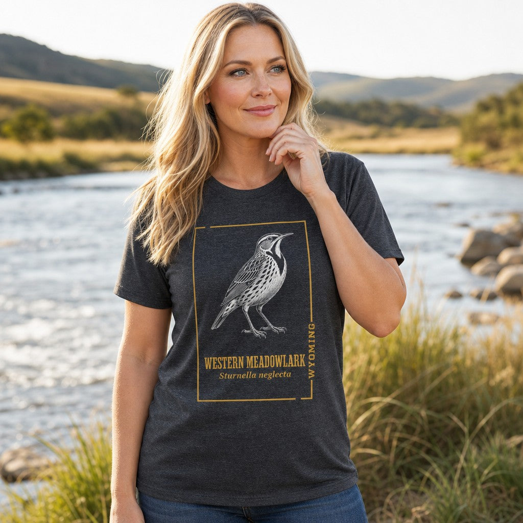 Woman in dark grey heather t-shirt featuring a vintage line art western meadowlark bird design with scientific name, standing by a scenic river, nature lovers, outdoors, by Crossroads Dandelion