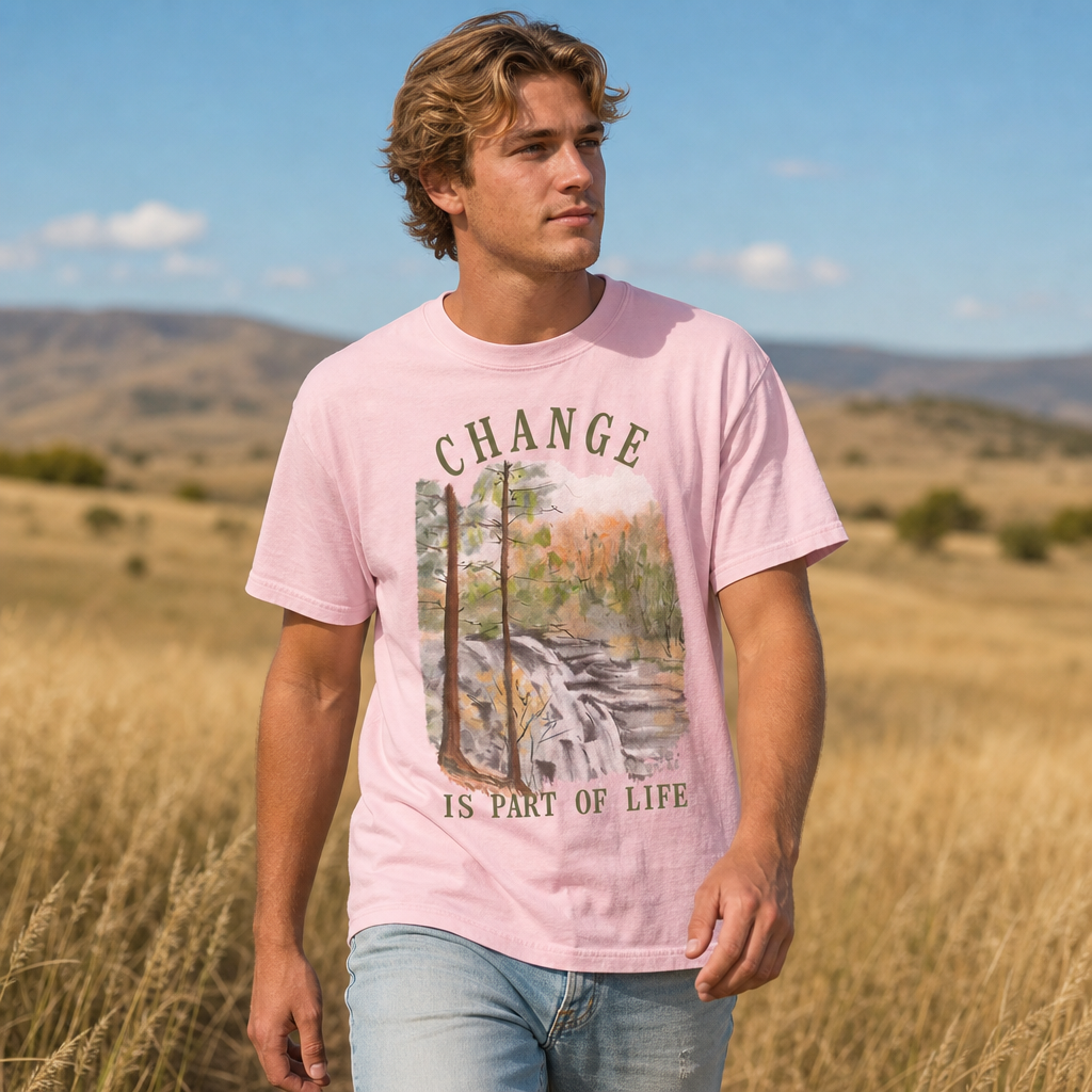 Man wearing blossom Comfort Colors 1717 t-shirt with watercolor forest and waterfall design and 'Change is part of life' text, standing in an open grassy field with mountains in the background, by Crossroads Dandelion