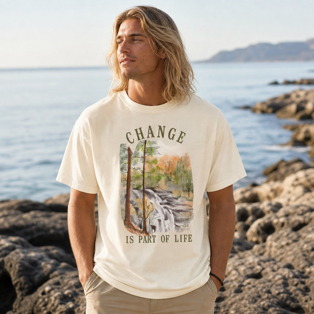 Man wearing ivory Comfort Colors t-shirt featuring a tranquil watercolor forest waterfall design with the phrase 'Change is part of life', standing by rocky beach with ocean view, by Crossroads Dandelion