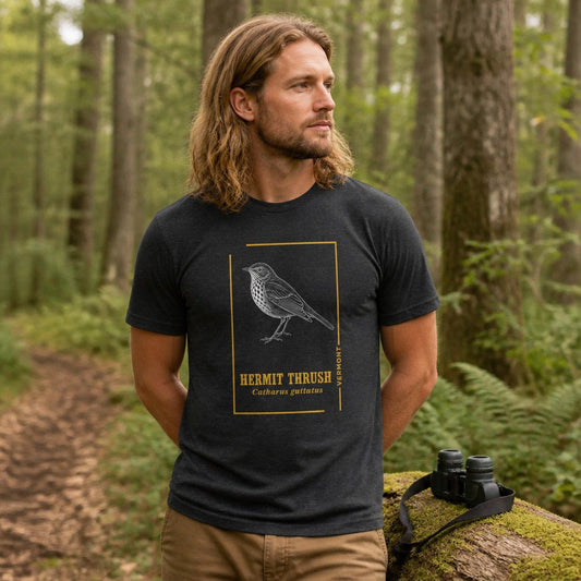 Man in dark grey heather Bella Canvas 3001 t-shirt featuring a vintage-style minimalist hermit thrush bird illustration with botanical details, birdwatching in a lush green forest, outdoors, perfect for nature lovers, men, and bird enthusiasts, by Crossroads Dandelion