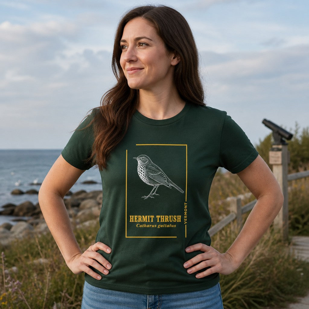 Woman in forest green Bella Canvas 3001 t-shirt featuring a vintage-style line art illustration of a hermit thrush with botanical details, minimalist folk-inspired design, standing on a coastal boardwalk with ocean view, perfect for birdwatchers and Vermont nature lovers, by Crossroads Dandelion