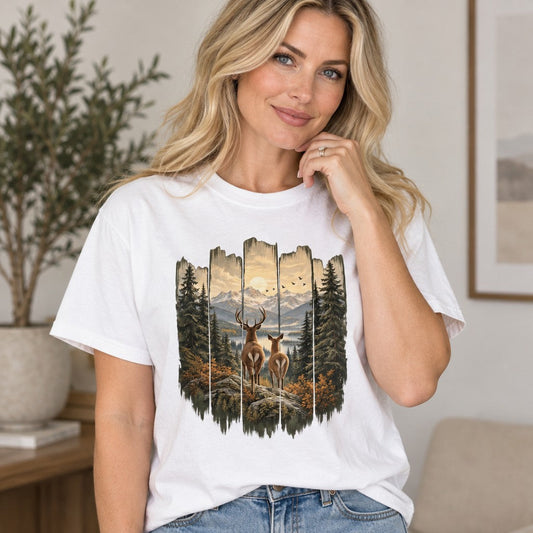 Woman in white Comfort Colors® 1717 t-shirt featuring a painterly folk-art wilderness design with a stag and doe on a rocky ledge, flanked by tall pines and golden autumn foliage, overlooking a valley with snow-capped mountains and birds in flight, rustic outdoor woodland style for nature enthusiasts and women, by Crossroads Dandelion
