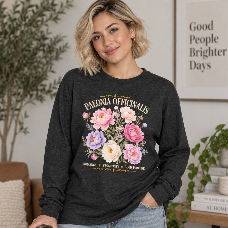 Woman wearing a dark grey heather Bella Canvas 3501 long sleeve shirt with a romantic watercolor peony and wildflower design in blush, cream, and violet hues, featuring vintage botanical script, cottagecore and garden-inspired, cozy living room setting, perfect for botanical enthusiasts and women, by Crossroads Dandelion