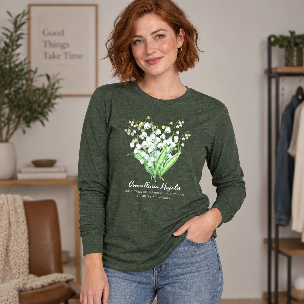 Red-haired woman in heather forest Bella Canvas 3501 long sleeve shirt featuring a vintage cottagecore watercolor Lily of the Valley botanical bouquet, standing in a cozy living room, ideal for floral illustration and botanical art lovers, by Crossroads Dandelion