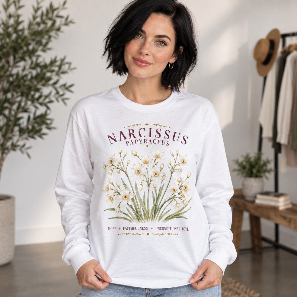 Woman wearing a white Bella Canvas 3501 long sleeve featuring a vintage watercolor narcissus floral design and ornate hand-lettered text, in a sunlit cottage-inspired living space, cottagecore and botanical lovers, by Crossroads Dandelion