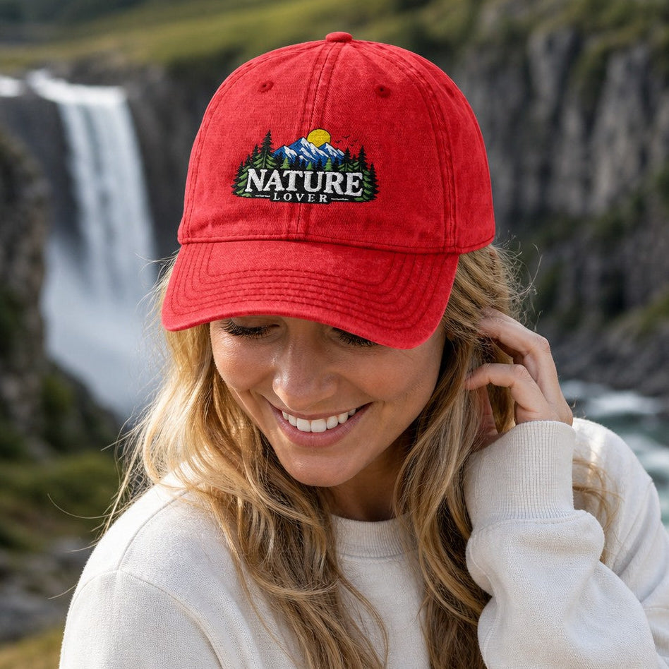 Woman wearing a red retro embroidered 'Nature Lover' baseball cap with mountain, forest, and sun design, smiling by a scenic waterfall, perfect for outdoor adventurers and nature lovers, by Crossroads Dandelion