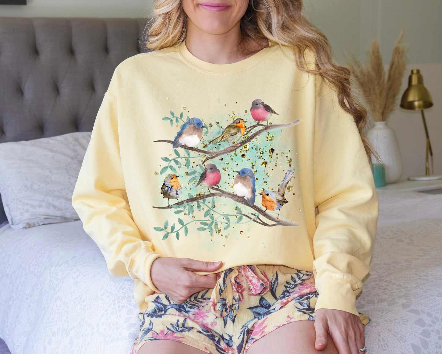 Woman wearing a Butter yellow sweatshirt with a watercolor style bird design sitting on a bed.
