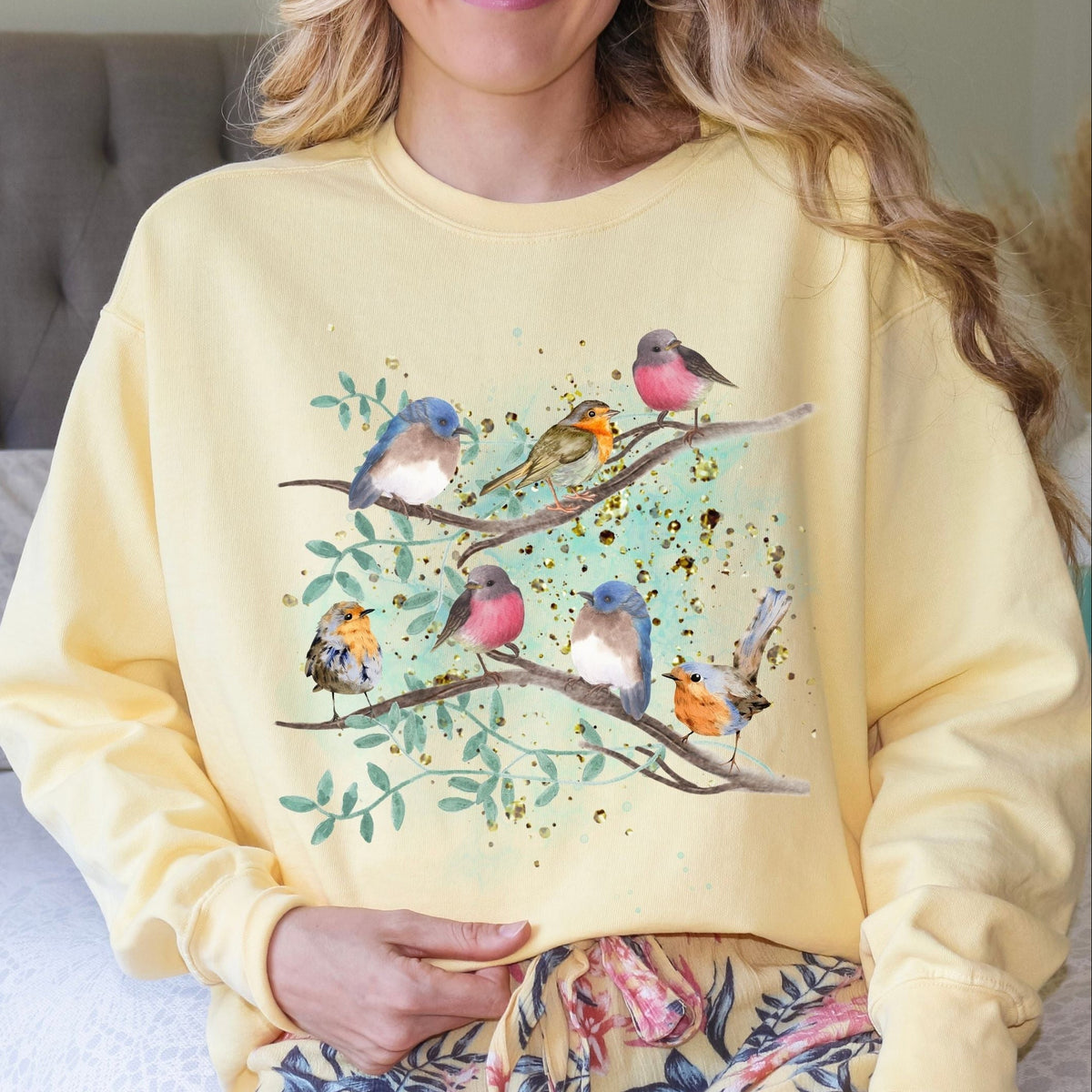 Woman wearing a Butter yellow sweatshirt with a watercolor style bird design sitting on a bed.