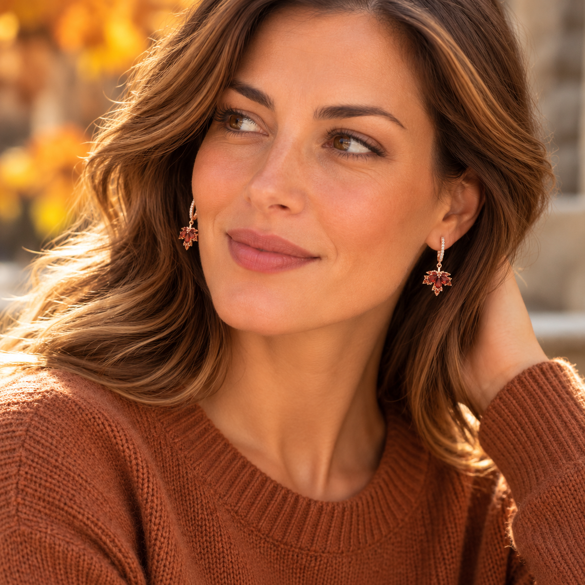 Woman wearing red maple leaf drop earrings and a rust-colored knit sweater against a softly blurred autumn background
