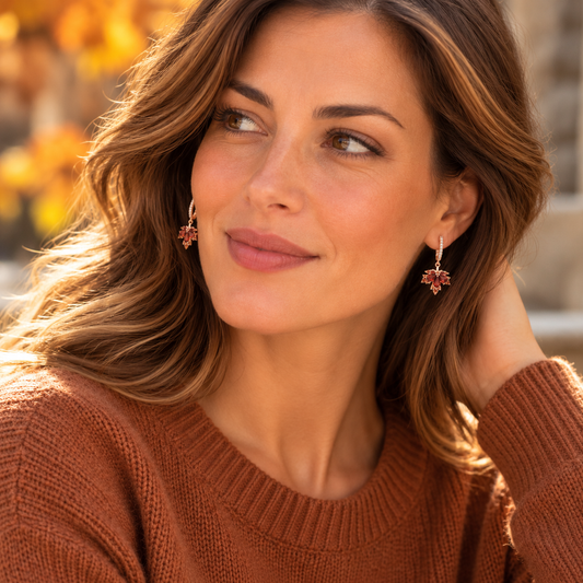 Woman wearing red maple leaf drop earrings and a rust-colored knit sweater against a softly blurred autumn background