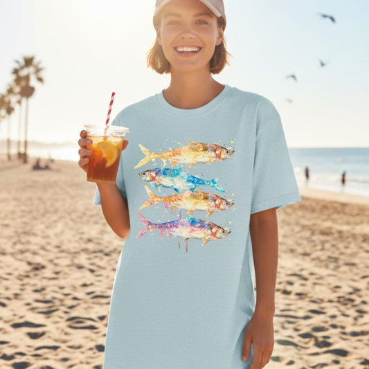 Woman on a beach wearing an oversized Heather Ice blue tee with colorful fish design, holding a drink. Retro watercolor sardines print for ocean lovers — Crossroads Dandelion