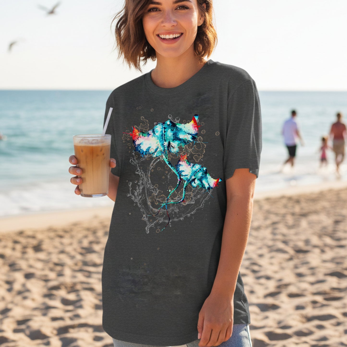 Woman on a beach holding an iced drink, wearing a dark grey heather t-shirt with a watercolor stingray graphic design.