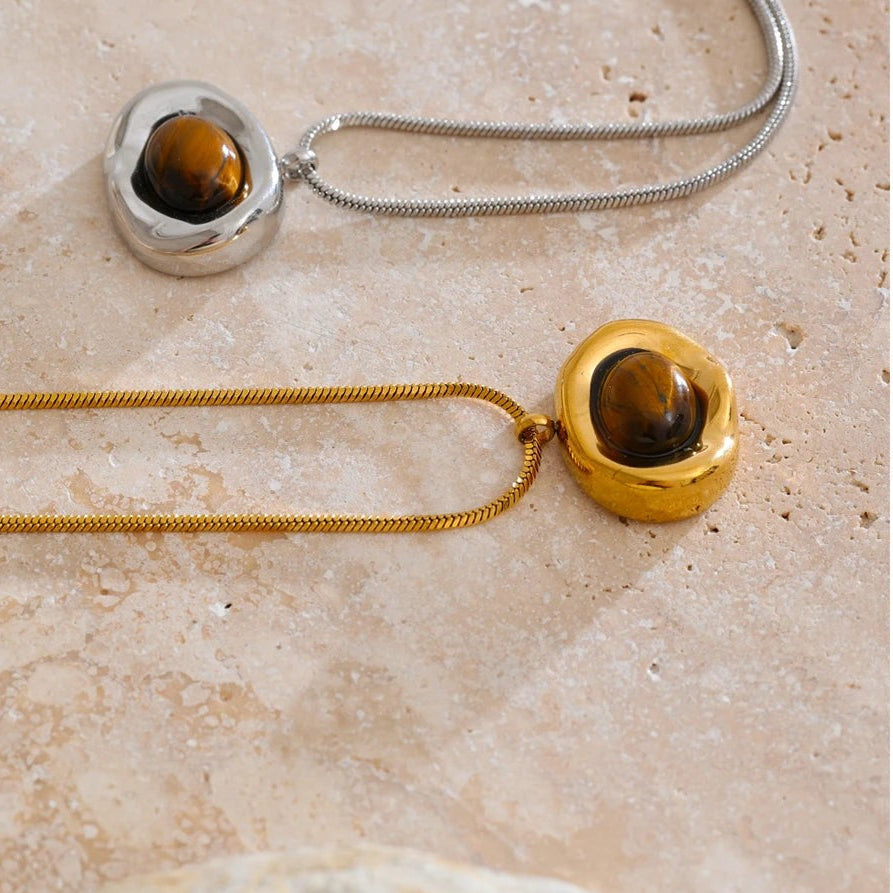Gold and silver tiger eye pendant necklaces laid flat on warm beige stone