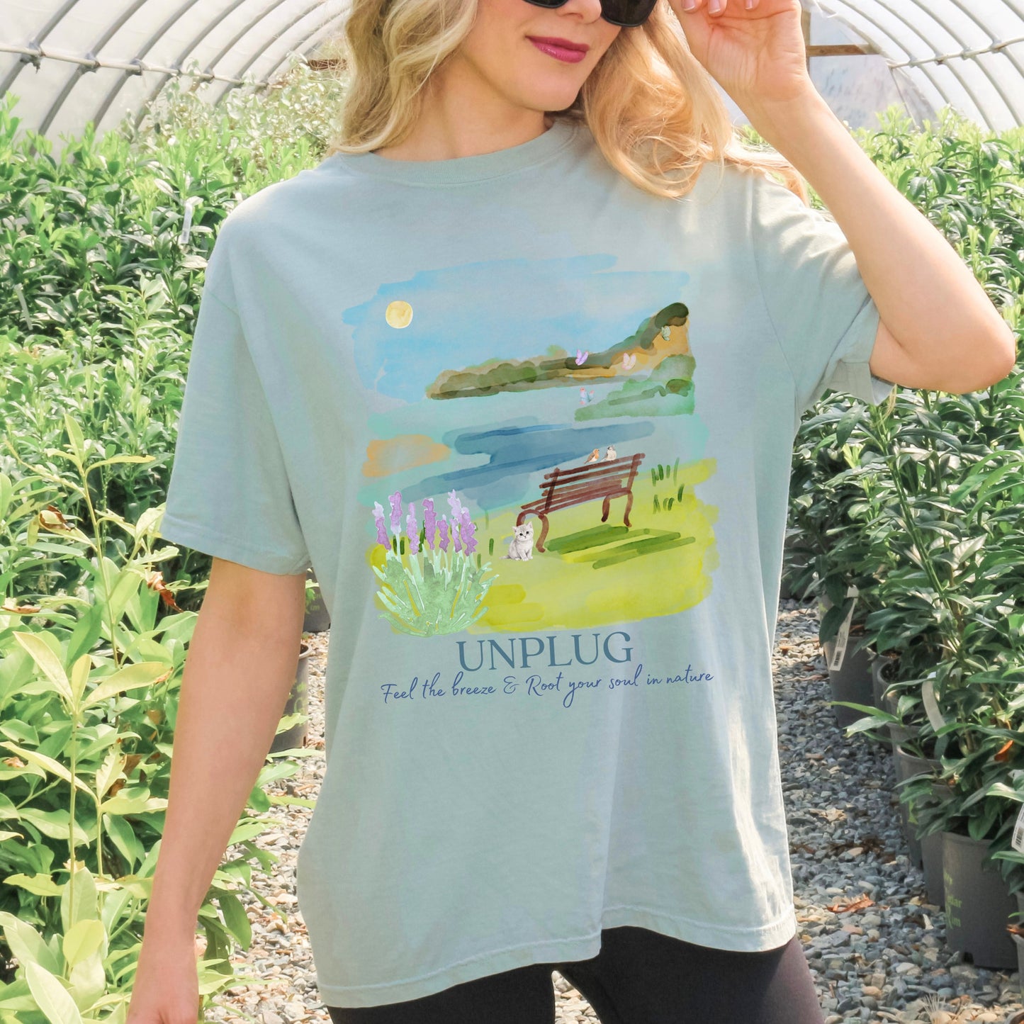 Woman wearing a bay color dyed tee Comfort Colors t-shirt with a watercolor mountain nature graphic and 'UNPLUG' text standing in a garden by Crossroads Dandelion