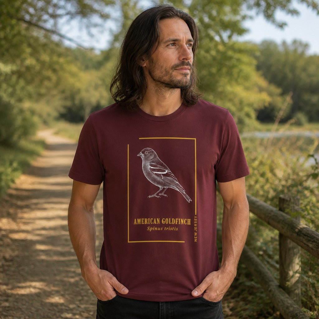 Man wearing a maroon Bella Canvas 3001 t-shirt with a vintage etching of an American Goldfinch framed in gold and classic serif text, on a nature trail, heritage birdwatching graphic for men and bird lovers, by Crossroads Dandelion