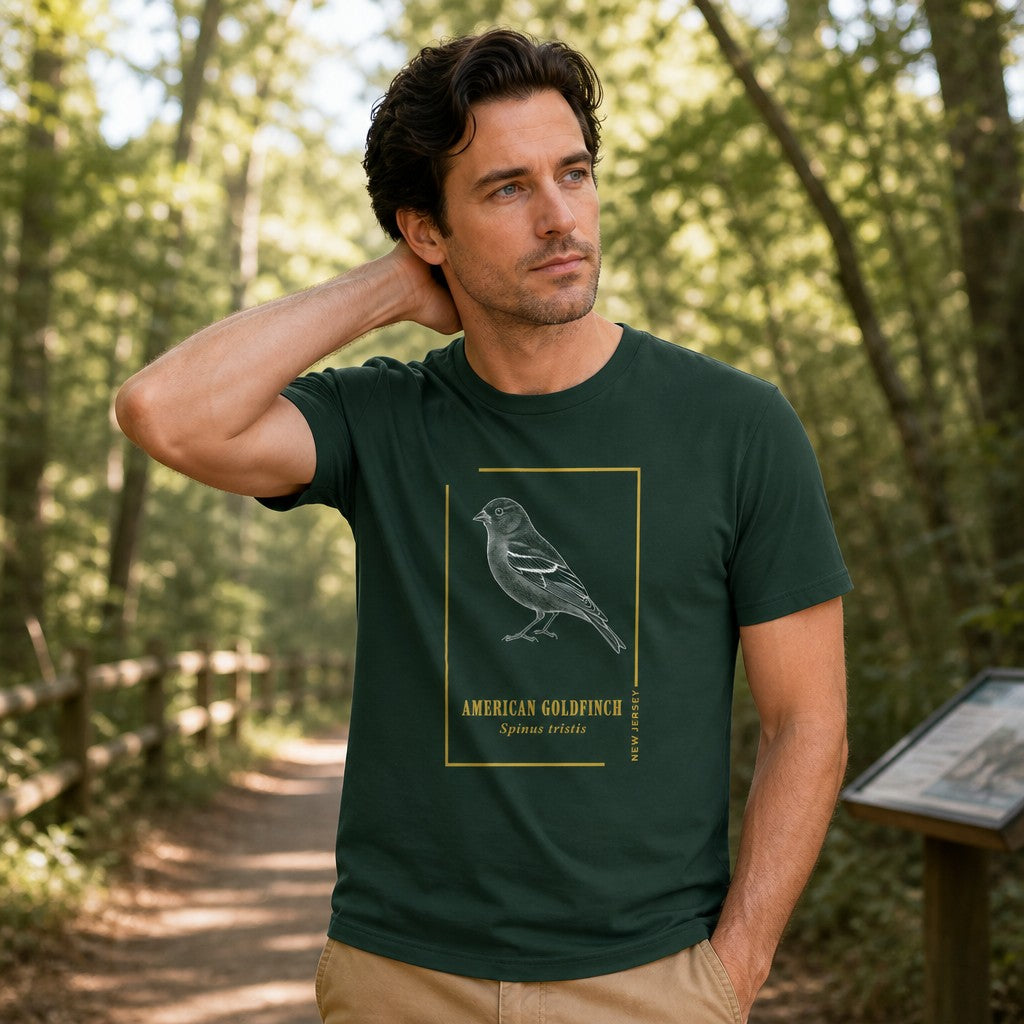 Man in forest green Bella Canvas 3001 t-shirt featuring a vintage etching botanical illustration of an American Goldfinch in a golden frame with serif lettering, classic field guide style, birdwatcher and naturalist gift for men, modeled on a wooded hiking trail, by Crossroads Dandelion