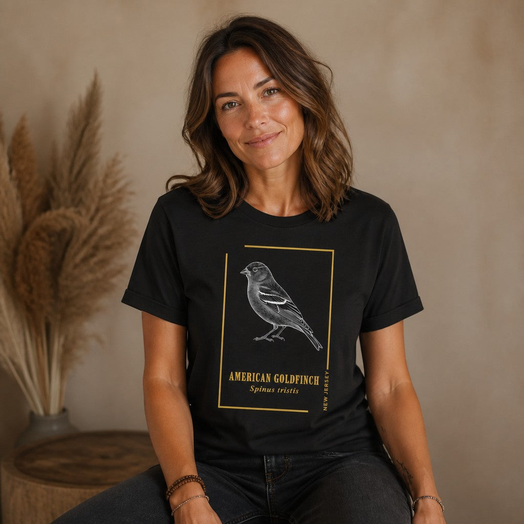 Woman wearing a black Bella Canvas 3001 shirt featuring a vintage etching illustration of an American Goldfinch in a golden frame with botanical serif lettering, classic field guide style, casual indoor setting, for birdwatchers, naturalists, and heritage wildlife art enthusiasts, by Crossroads Dandelion