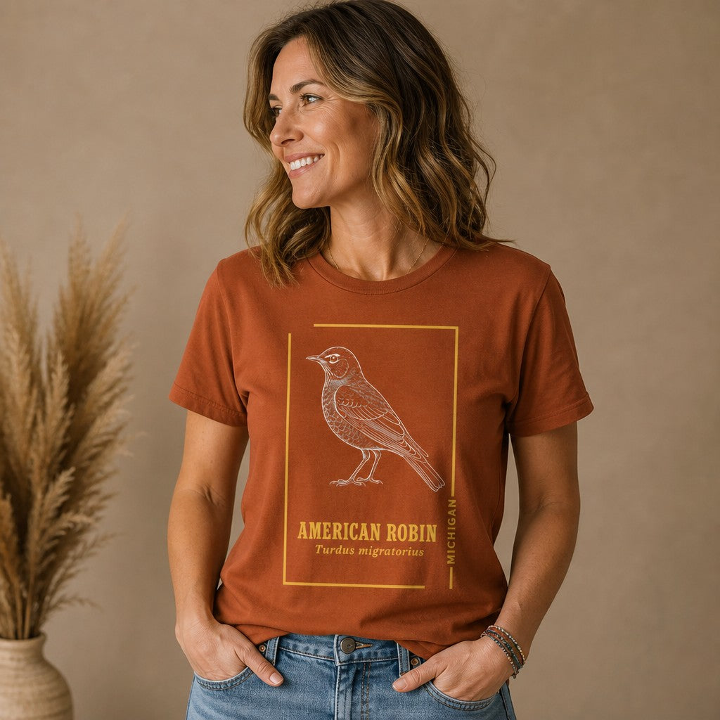 Woman wearing an autumn-colored Bella Canvas 3001 t-shirt featuring a vintage field-guide style white line art American robin illustration with minimalist gold border and elegant typography, designed for birdwatchers and cottagecore enthusiasts, by Crossroads Dandelion