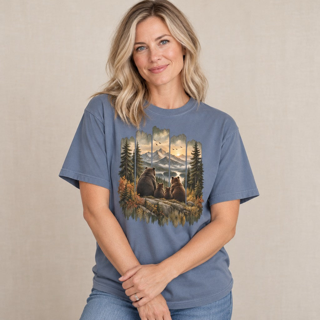 Woman in blue jean Comfort Colors® 1717 t-shirt featuring a vintage-style painted scene of a brown bear family sitting on a rocky ledge, overlooking snow-capped mountains and a sunset river, with distressed wood-panel framing, rustic cabin art style, for hikers, campers, and wildlife lovers, by Crossroads Dandelion