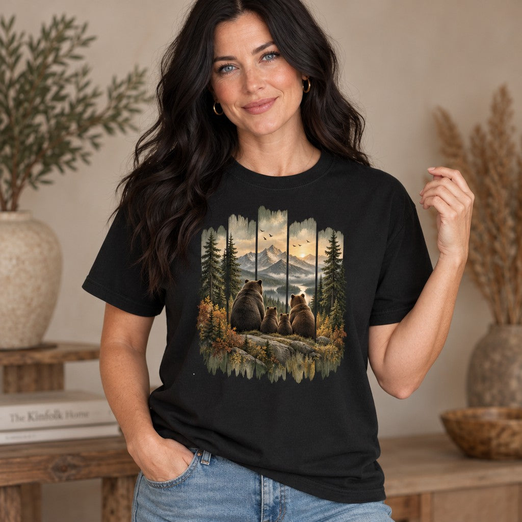 Woman wearing black Comfort Colors® t-shirt featuring a vintage-style painted scene of a brown bear family on a rocky ledge overlooking snow-capped mountains, a winding river, golden sunset, and evergreen woods, framed by distressed wood for a rustic, cabin-inspired look; perfect for hikers, campers, and wildlife lovers, by Crossroads Dandelion