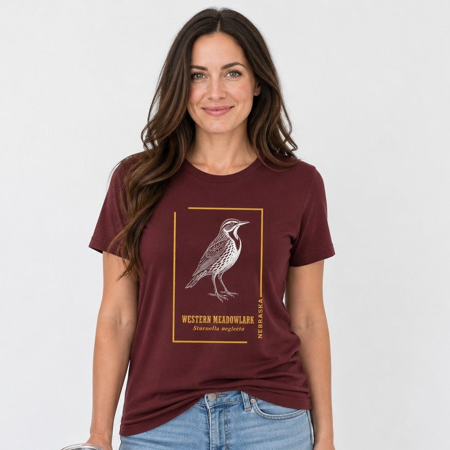 Woman in maroon Bella Canvas 3001 t-shirt featuring a vintage-inspired white line art Western Meadowlark bird illustration with gold frame and botanical typography, classic field guide nostalgic style, casual setting, perfect for birders and nature lovers, by Crossroads Dandelion
