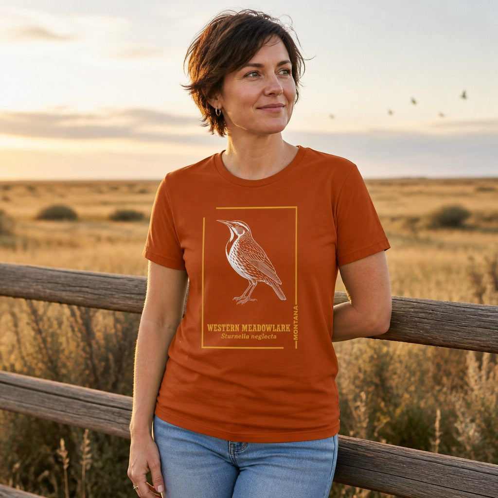 Woman wearing an autumn orange Bella Canvas 3001 t-shirt featuring a vintage hand-drawn etching of a Western Meadowlark in white with golden frame and field guide typography, standing by a wooden fence in a prairie setting, birdwatcher gift, heritage nature art, by Crossroads Dandelion