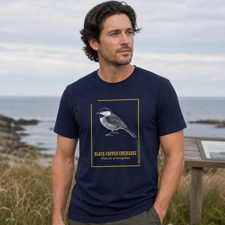 Man wearing navy Bella Canvas 3001 t-shirt with vintage naturalist engraving of a black-capped chickadee, golden line frame, and scientific text, standing by the coast, for bird lovers and cottagecore wildlife art enthusiasts, by Crossroads Dandelion