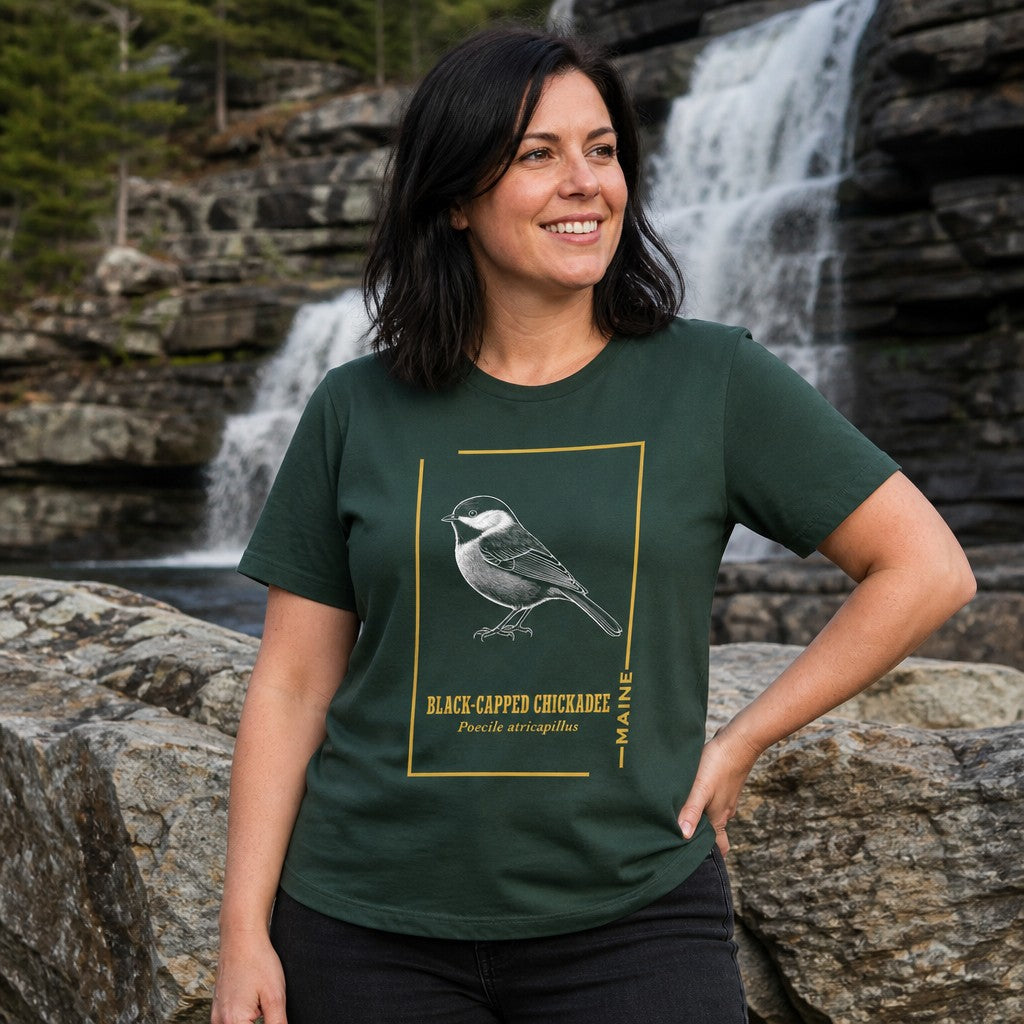 Woman wearing a forest green t-shirt featuring a vintage line art black-capped chickadee bird illustration, standing near a waterfall, ideal for bird watchers and nature lovers, by Crossroads Dandelion